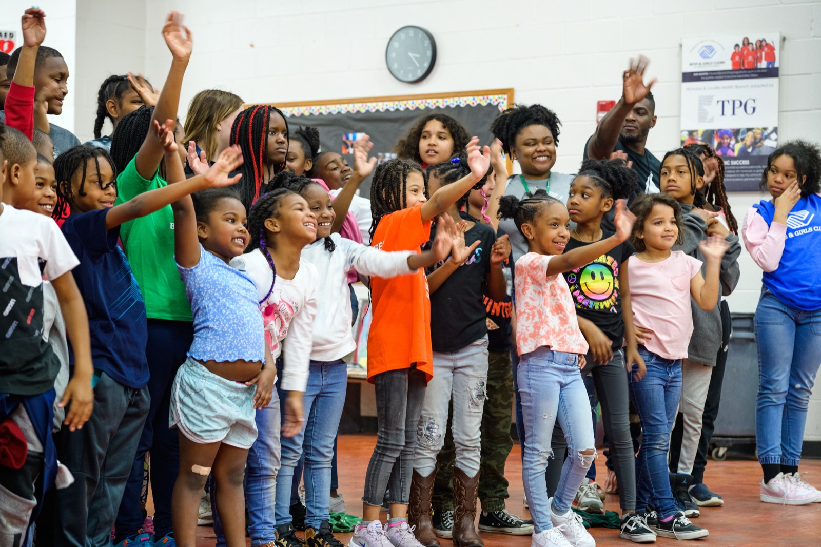 Group of children waving and smiling together in a gym, with Club staff standing behind them