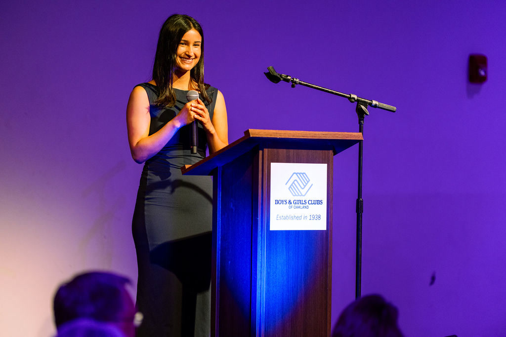A Youth of the Year speaker smiling as she holds a microphone at a podium with a Boys & Girls Clubs of Oakland sign