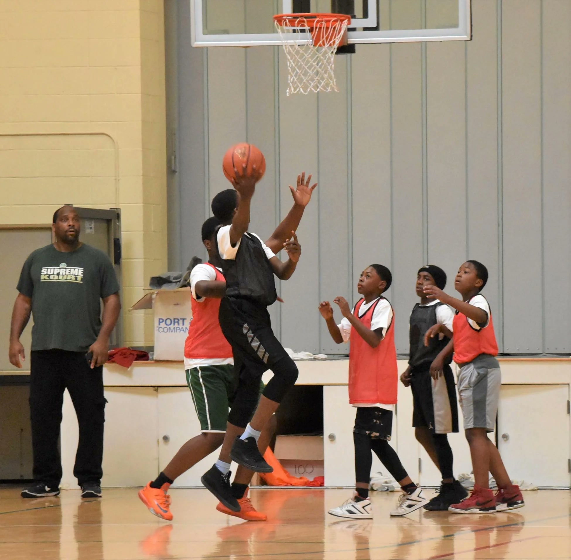 Boys playing a basketball game in a gym, one jumping to shoot as others and a coach look on