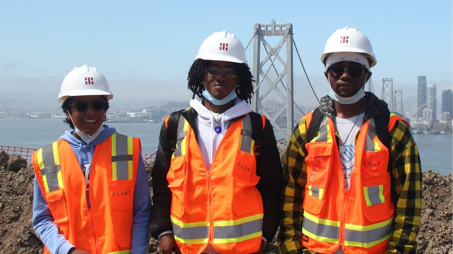 Three Club teens in hard hats and orange safety vests standing in front of the Bay Bridge