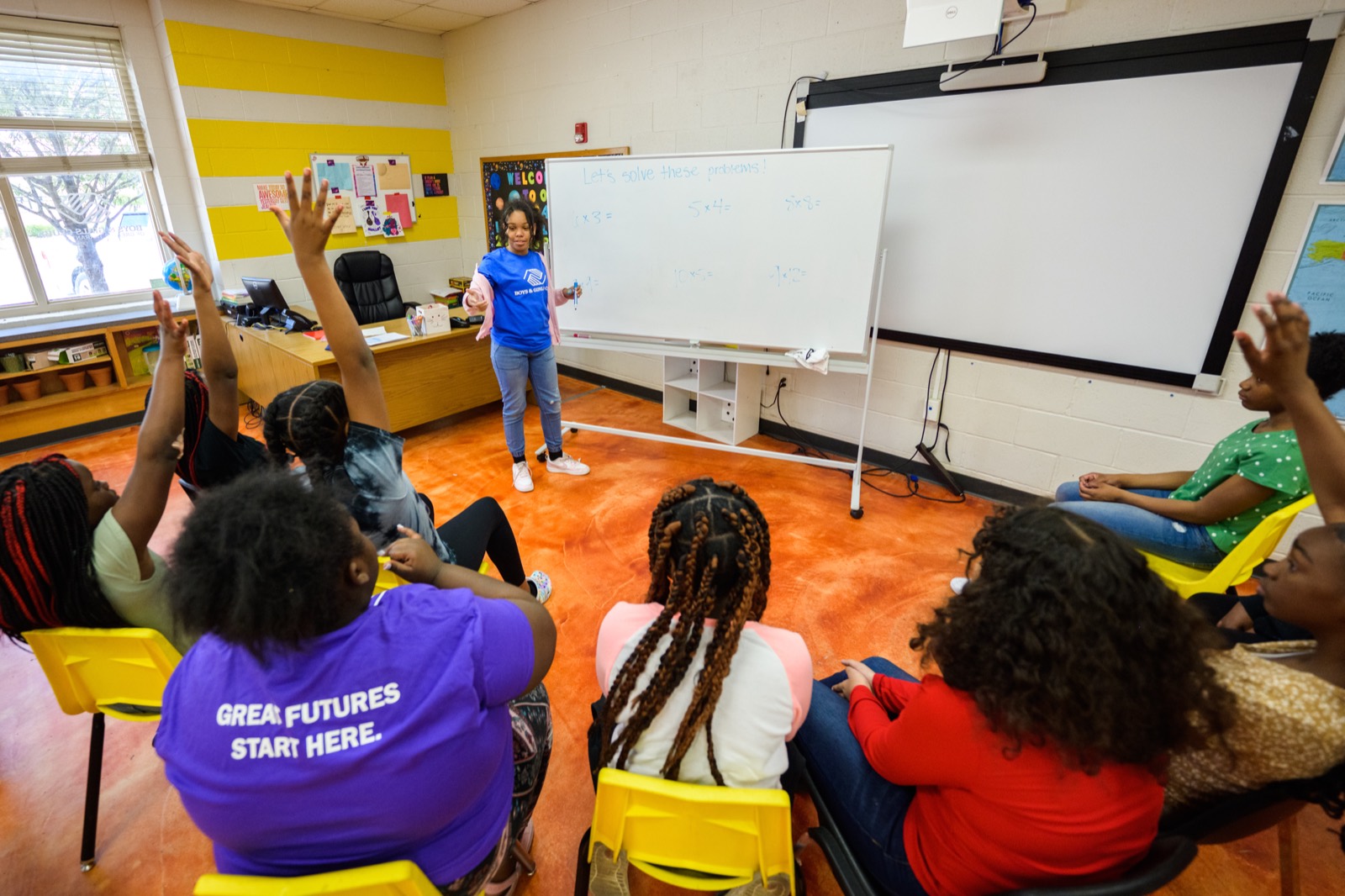 Teens raising their hands in a classroom as a young leader in a blue shirt teaches at a whiteboard