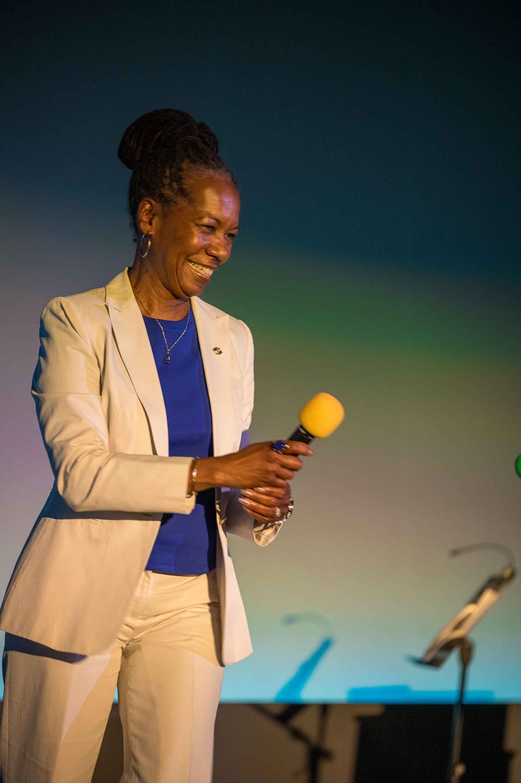 A woman in a cream blazer speaking into a yellow microphone on stage