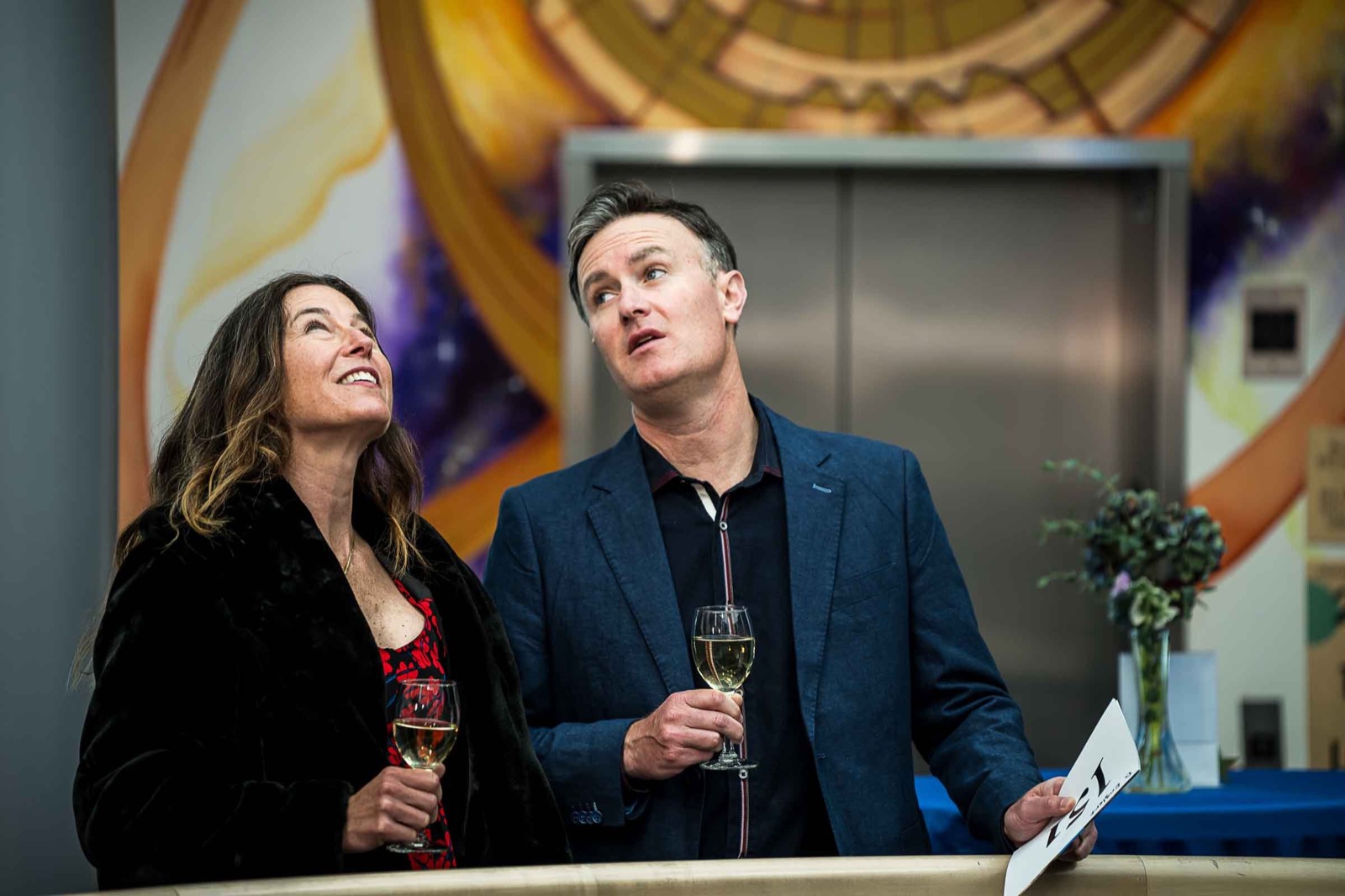 A woman and a man in a navy blazer holding drinks and looking up together at the gala