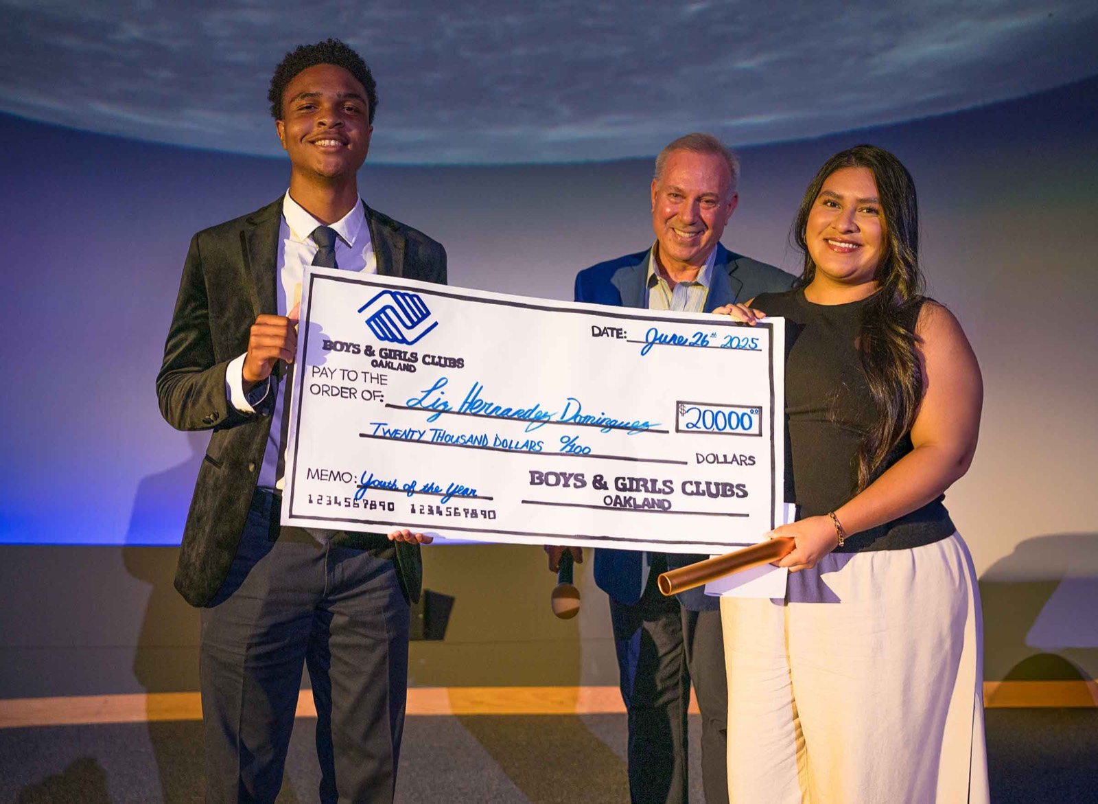 A Youth of the Year winner and two presenters holding a large Boys & Girls Clubs of Oakland check on stage