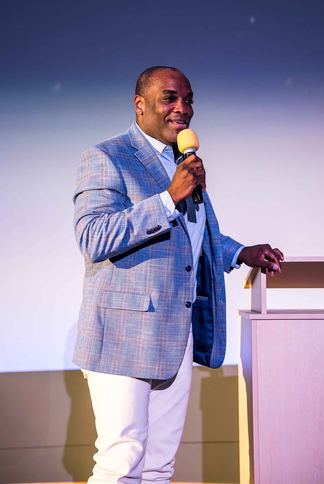 A man in a blue plaid blazer speaking into a yellow microphone on stage