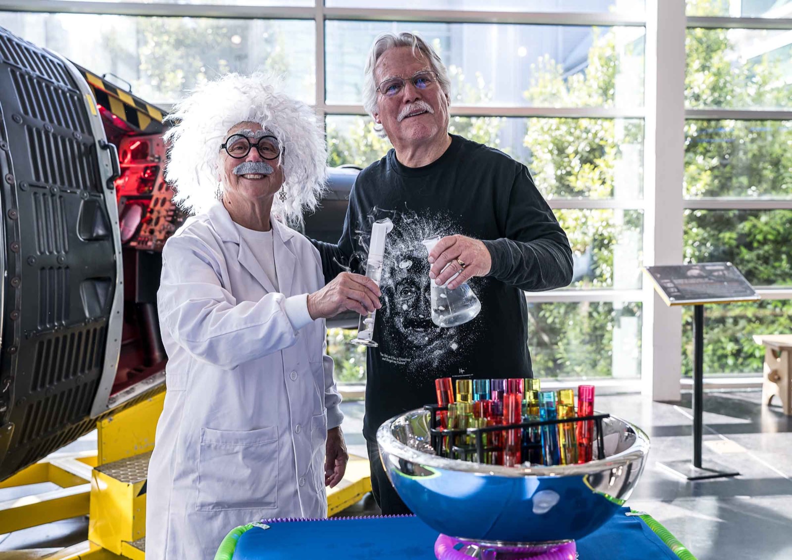 A guest in a lab coat and wild white wig costume and a man in black handling glass beakers beside a spacecraft capsule
