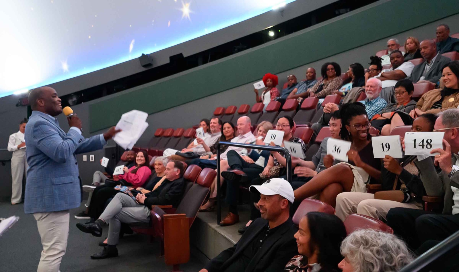 An emcee holding papers and a microphone in front of a seated audience in the planetarium theater, with auction paddles visible