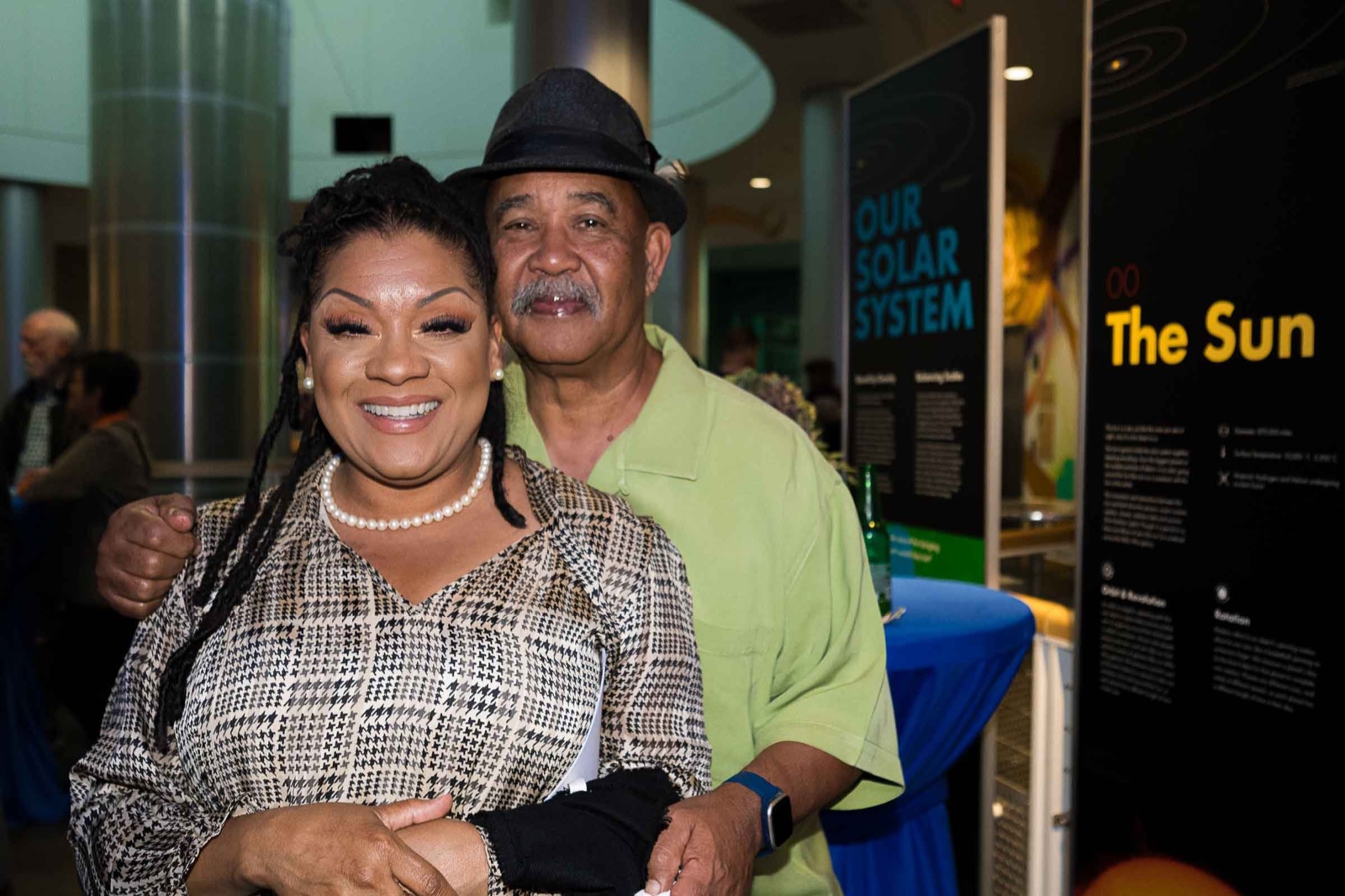 A smiling woman in a pearl necklace and an older man in a hat and green shirt posing in front of solar system exhibit panels