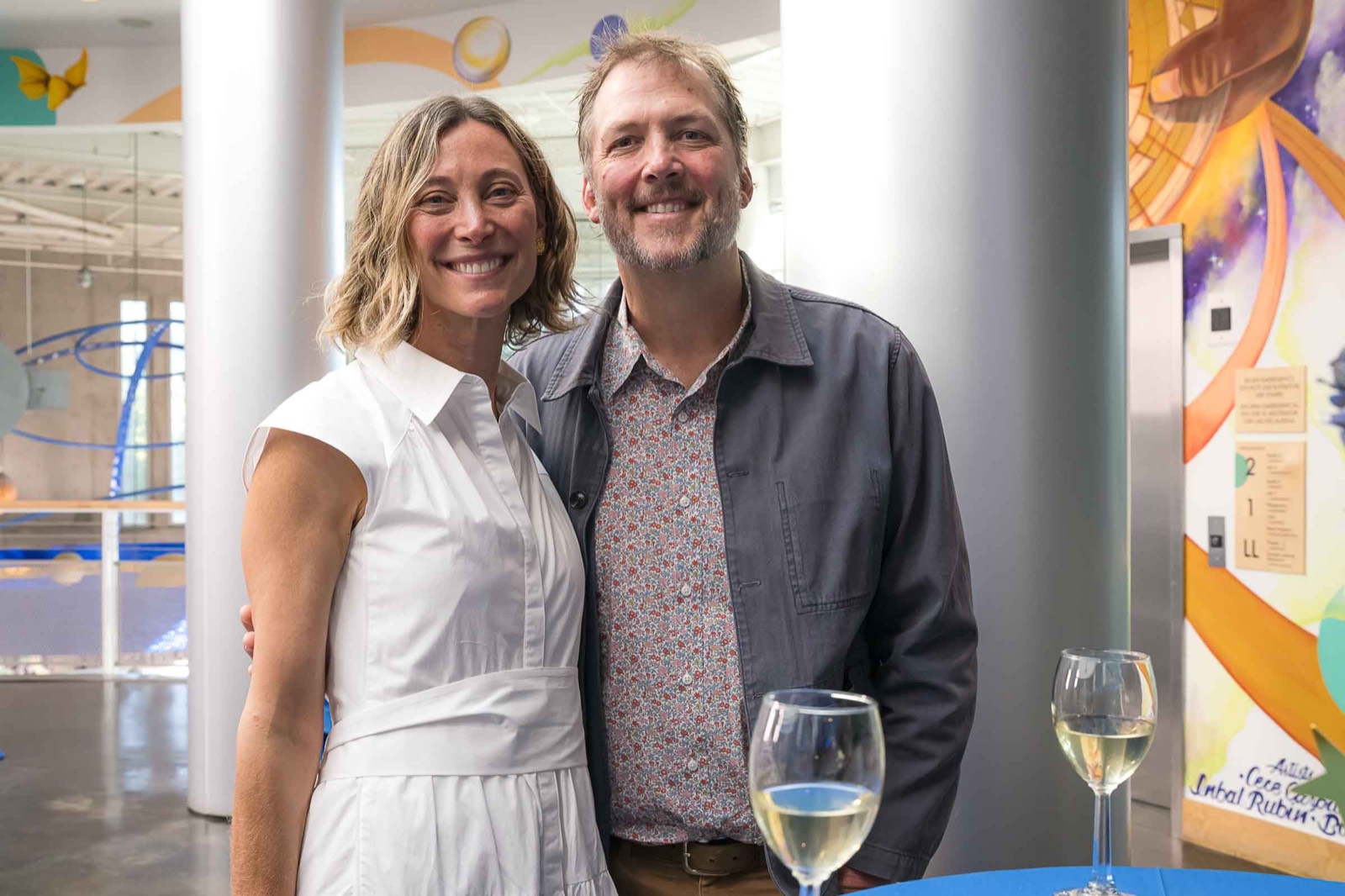 A smiling couple posing with glasses of wine in a bright atrium at the gala