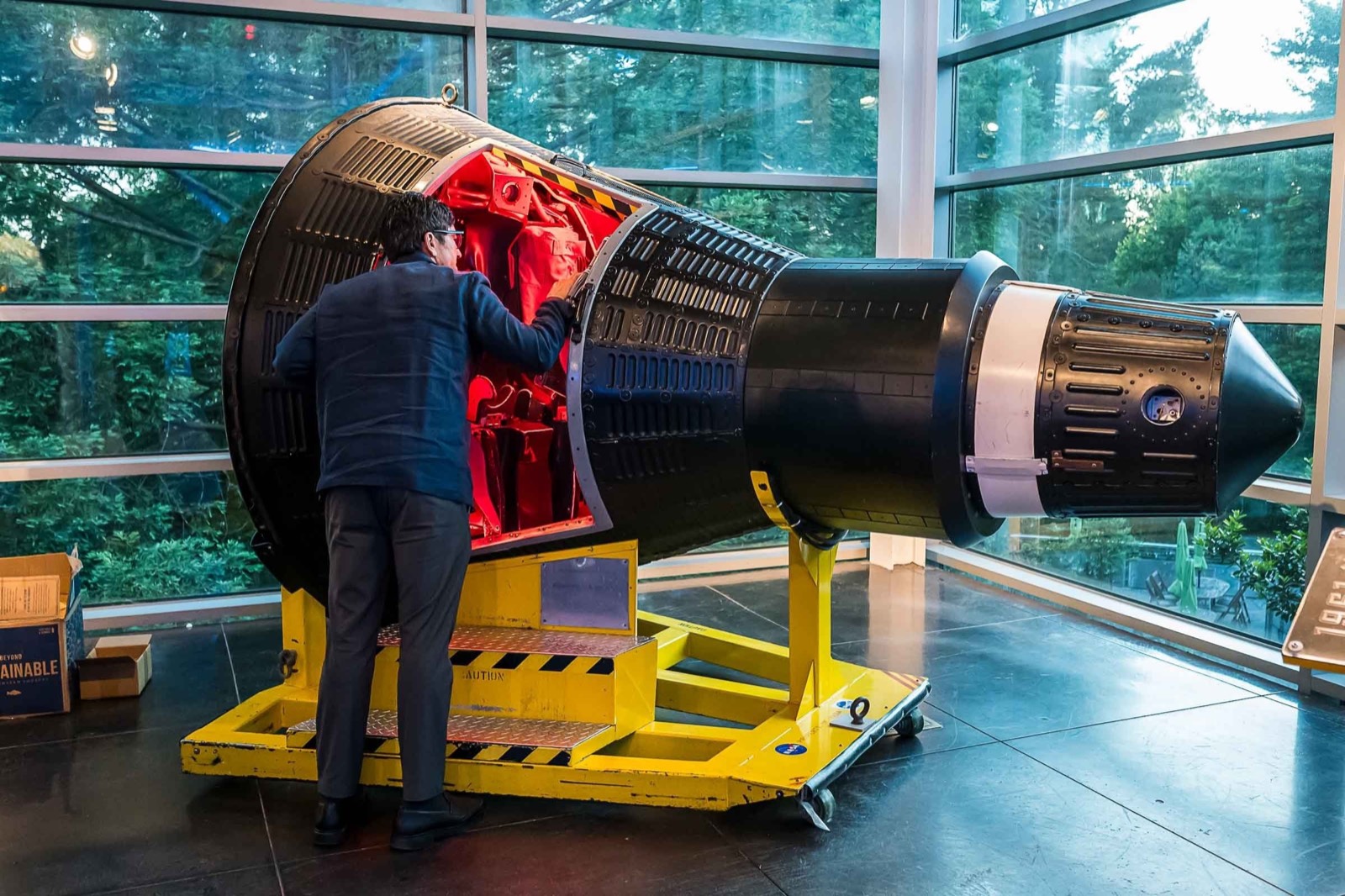 A man examining the red interior of a black spacecraft capsule on a yellow stand in a glass-walled hall