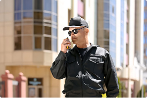 Security officer in a cap and sunglasses speaking into a handheld radio in front of a large building