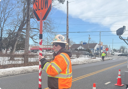 Flagger in an orange jacket and hard hat holding a SLOW sign on a road in winter