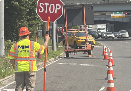 MJM Ultimate Services flagger in an orange hard hat holding a STOP sign beside traffic cones on a highway ramp