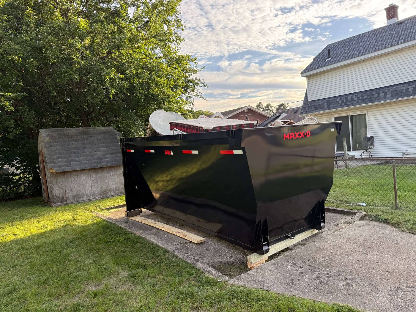 Large black Maxx-D dumpster placed on wooden planks on a concrete slab in a yard near a white house and a small shed, filled with old furniture and items.