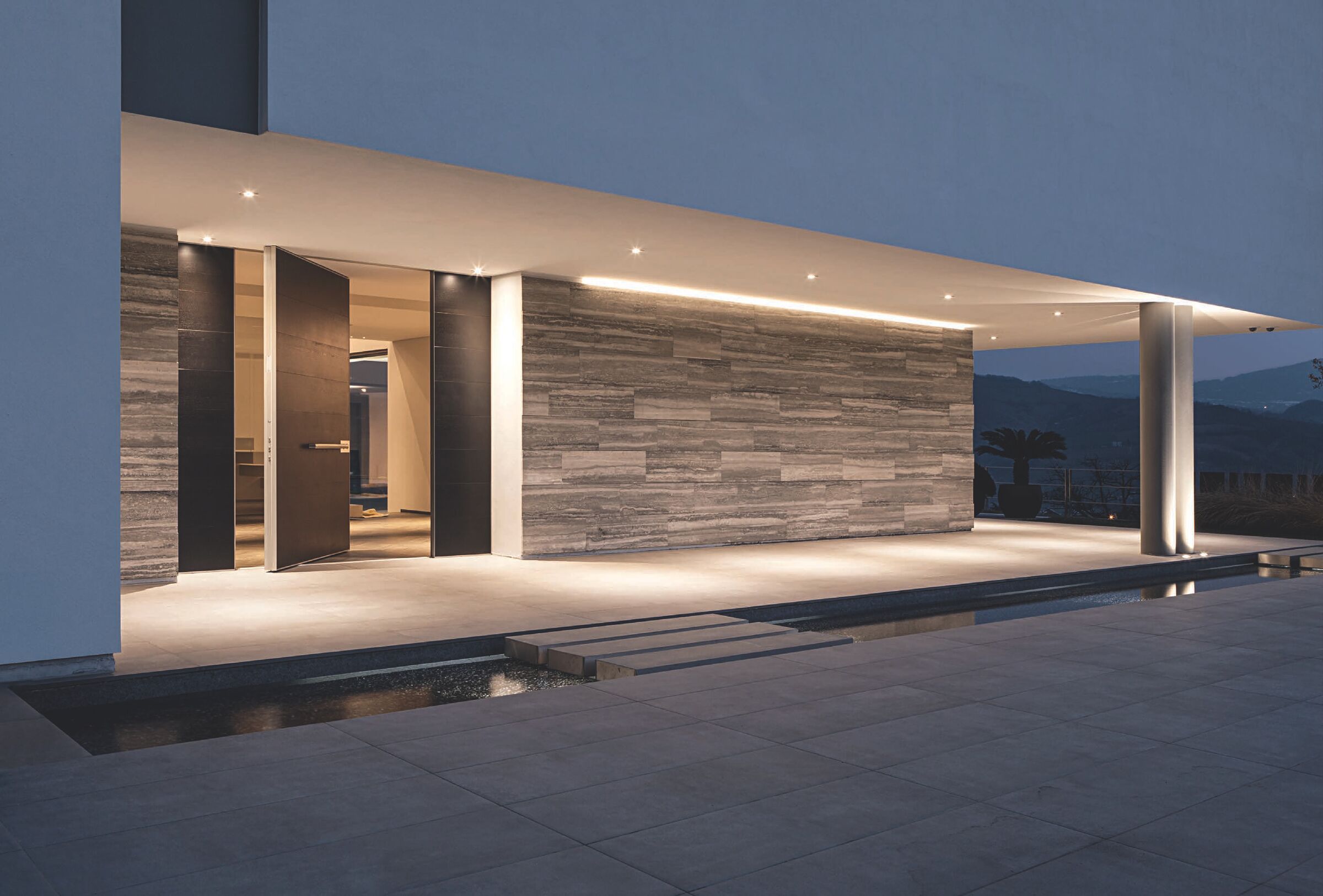 European-style pivot entrance door at dusk beside a stone wall and reflecting pool