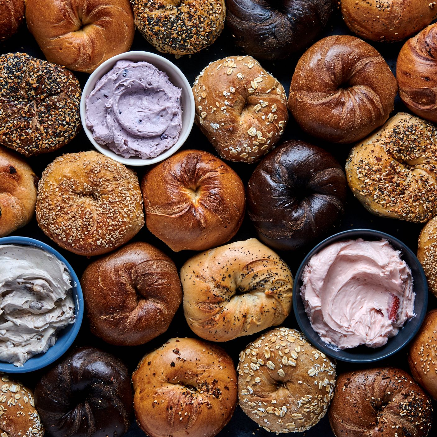 Assorted bagels with three bowls of flavored cream cheese on a dark surface.