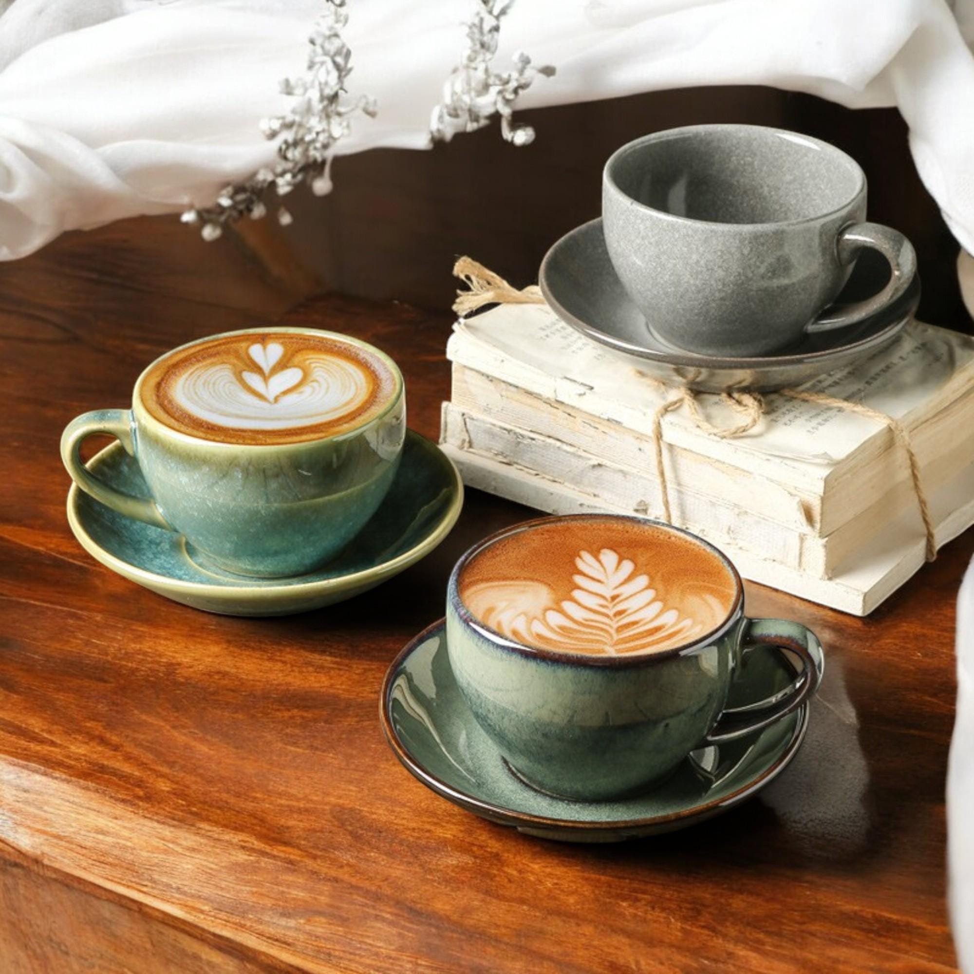 Two green cups of latte with leaf latte art on wooden table near stacked books with gray cup.