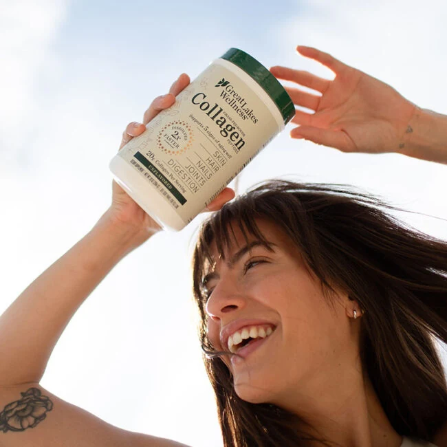 A smiling woman holding a jar of Great Lakes Wellness collagen above her head against a bright sky