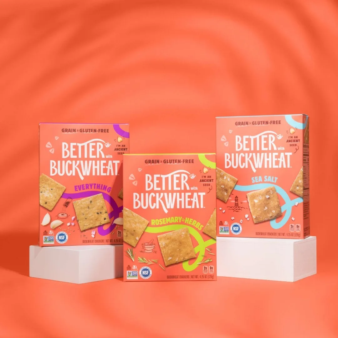 Three boxes of Better With Buckwheat crackers on white blocks against an orange background