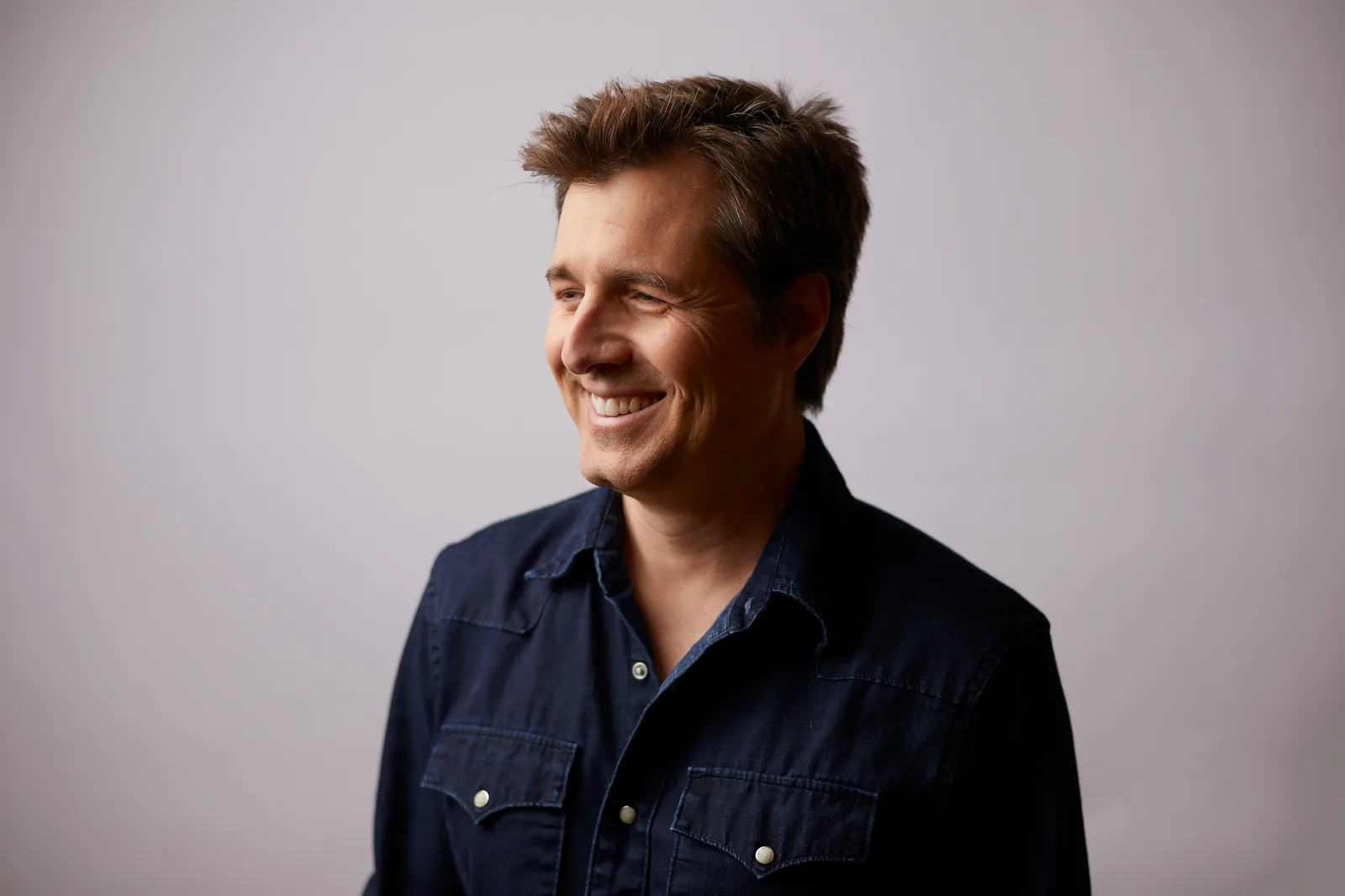 A smiling man in a dark denim shirt photographed against a grey studio backdrop