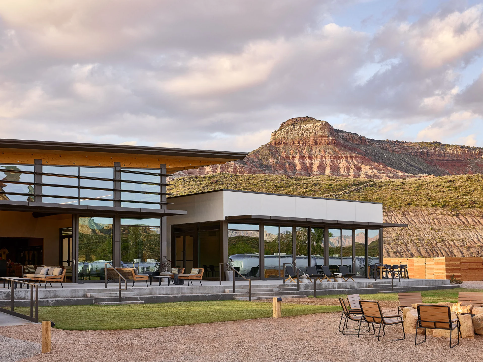 A glass-fronted Autocamp clubhouse at dusk with an Airstream behind the windows, a red rock mesa beyond and fire pit chairs in the foreground