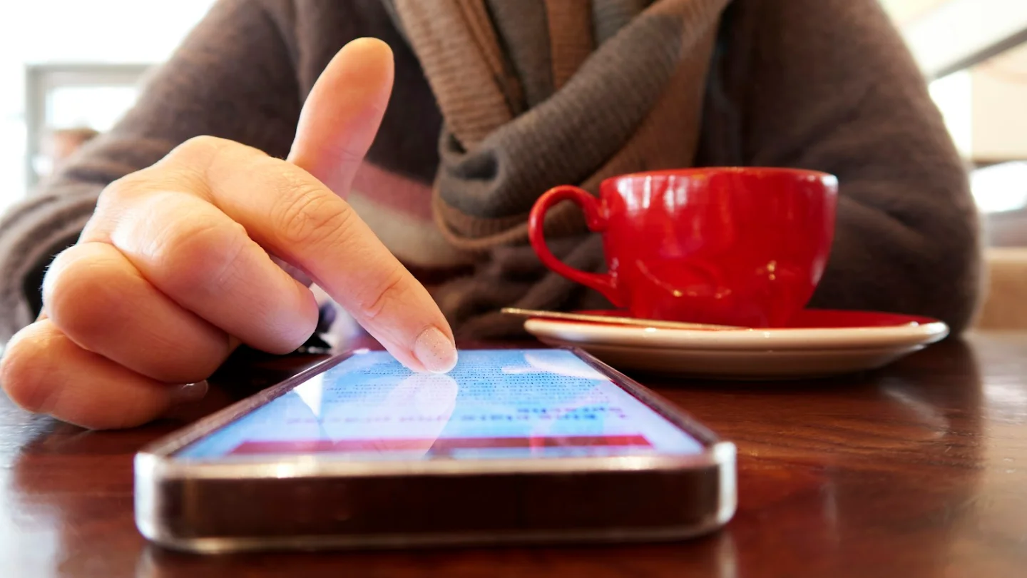 A person tapping a smartphone screen at a cafe table next to a red coffee cup