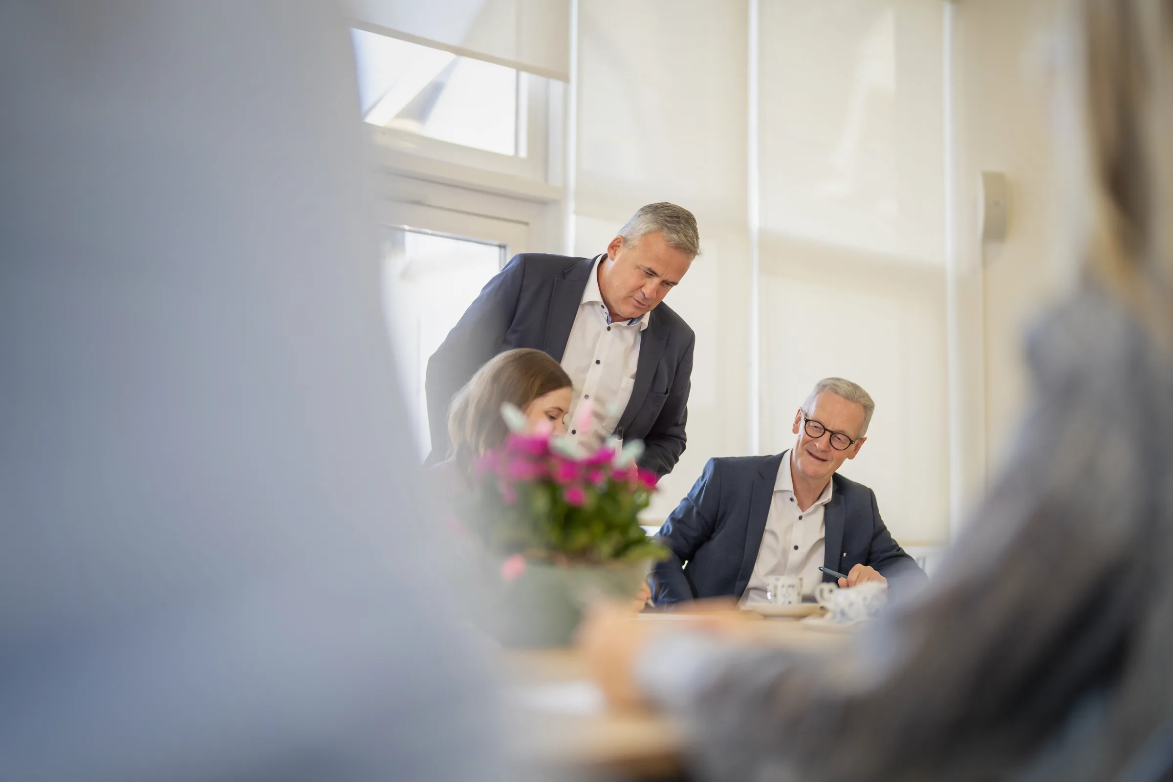 Drei Geschäftspersonen in einem modernen Büro bei einer Besprechung, zwei Männer in Anzügen und eine Frau, die konzentriert arbeitet, mit einer Pflanze und Teetassen auf dem Tisch.