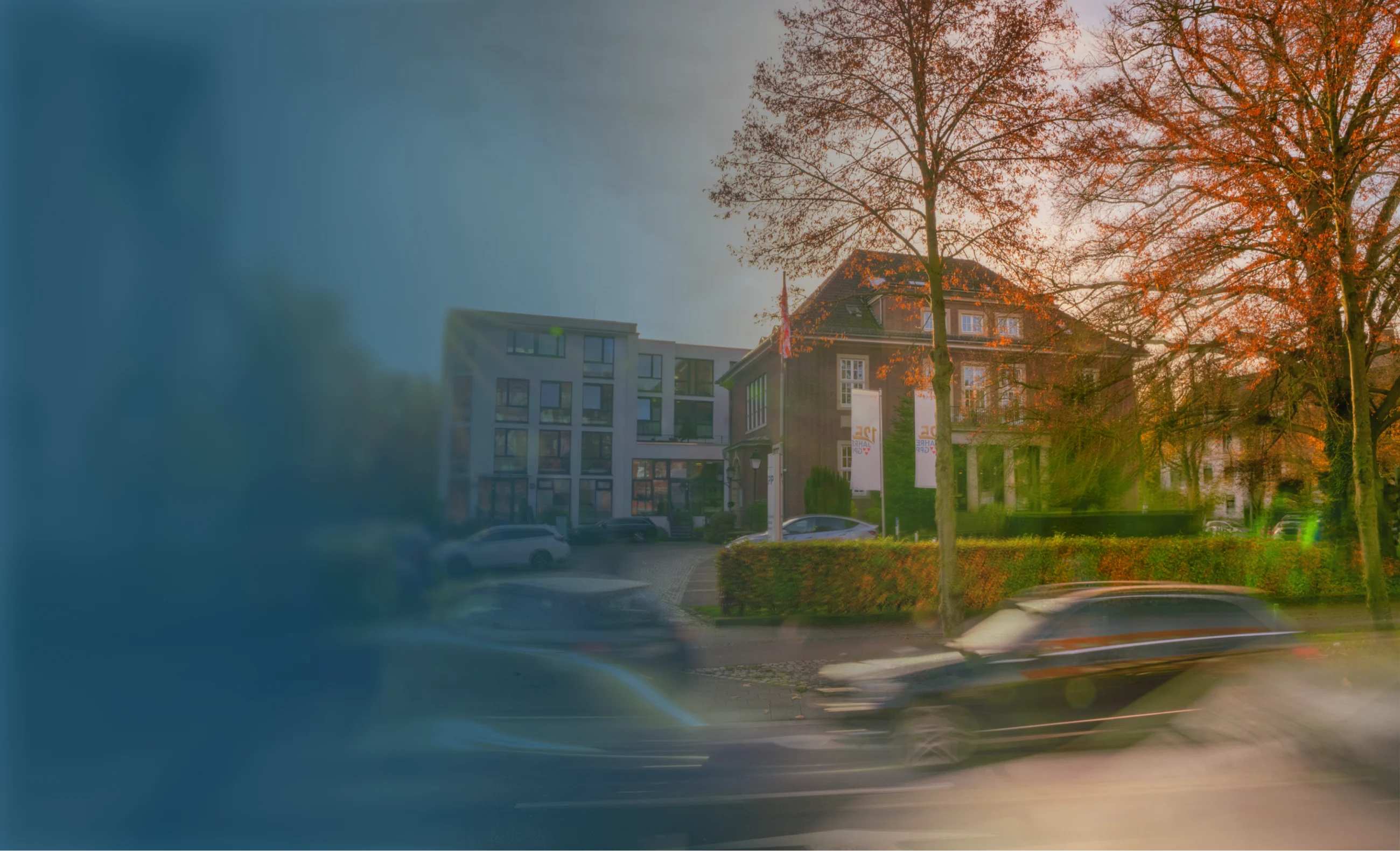 Straßenszene mit verschwommen fahrenden Autos vor einem historischen Gebäude neben moderner Architektur und Bäumen mit herbstlichen Blättern.
