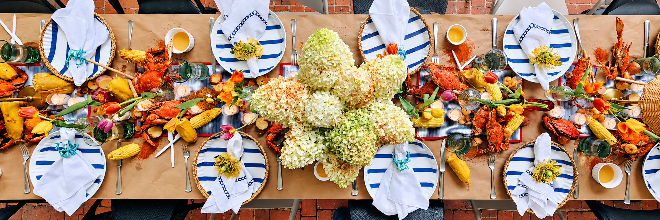 Overhead view of a colorful event table with seafood, corn, flowers and blue-striped place settings