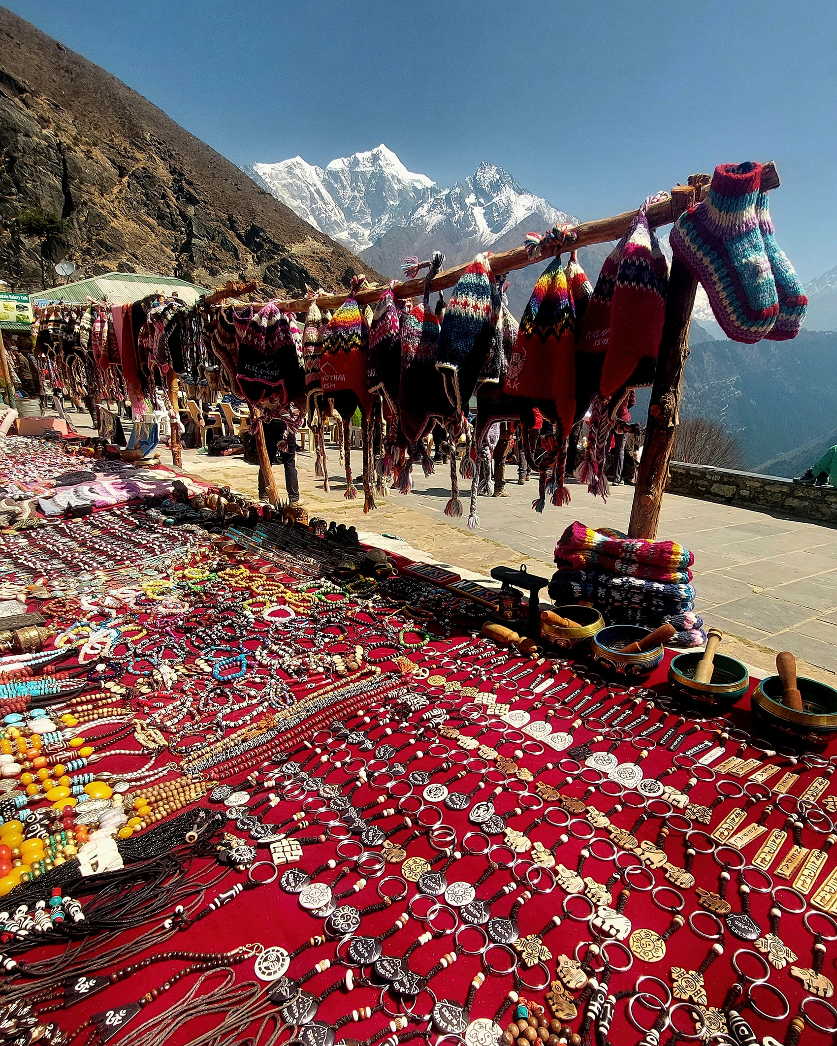 Colourful knitted hats, jewellery and handicrafts at an outdoor market in Nepal, with Himalayan mountains in the background.