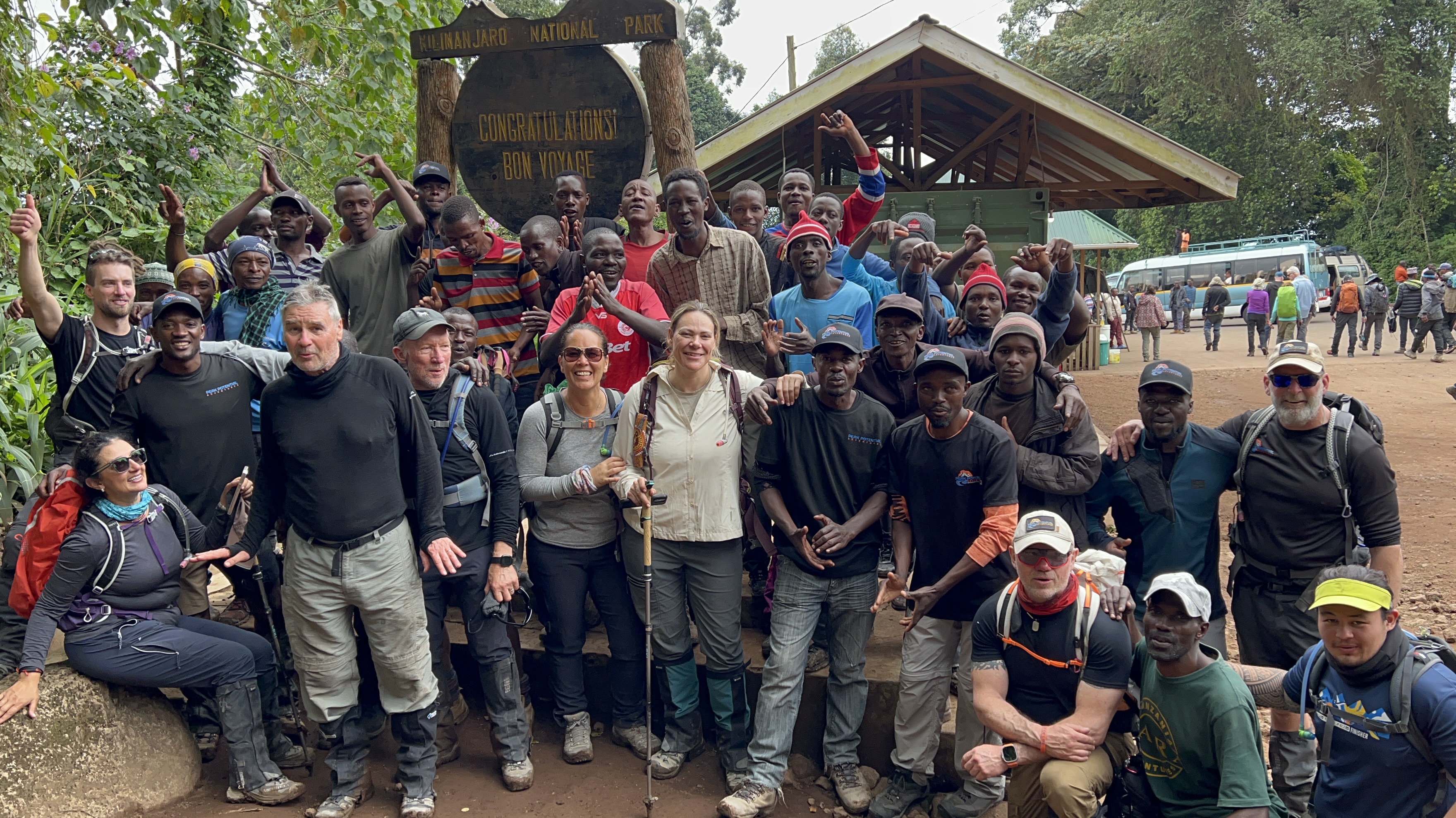 Peak Potential Adventures trekkers and local guides celebrating together beside the Kilimanjaro National Park sign in Tanzania.