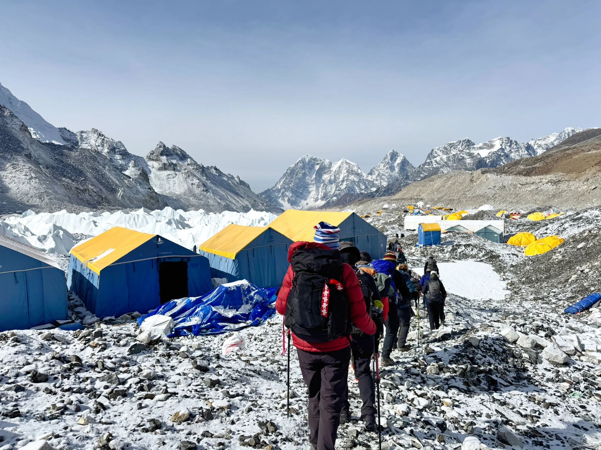 Trekkers crossing rocky, snow-covered ground near Everest Base Camp, with expedition tents and Himalayan mountains beyond.