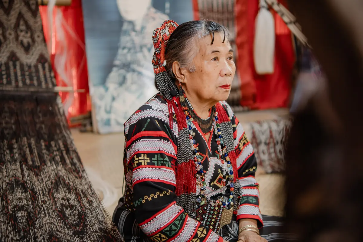 An elder weaver in Tboli dress and beaded headpiece, seated beside a loom