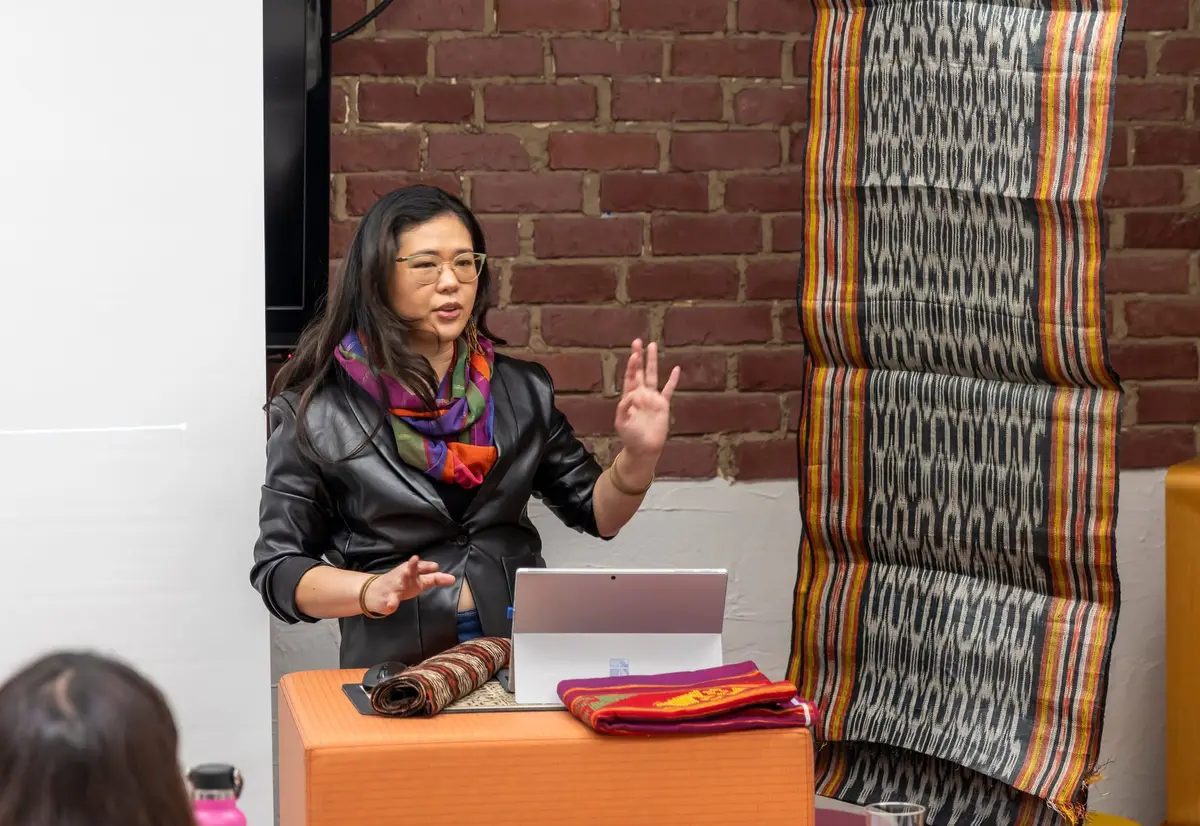 A speaker presenting beside a length of T’nalak hung on a brick wall, with folded textiles on the table