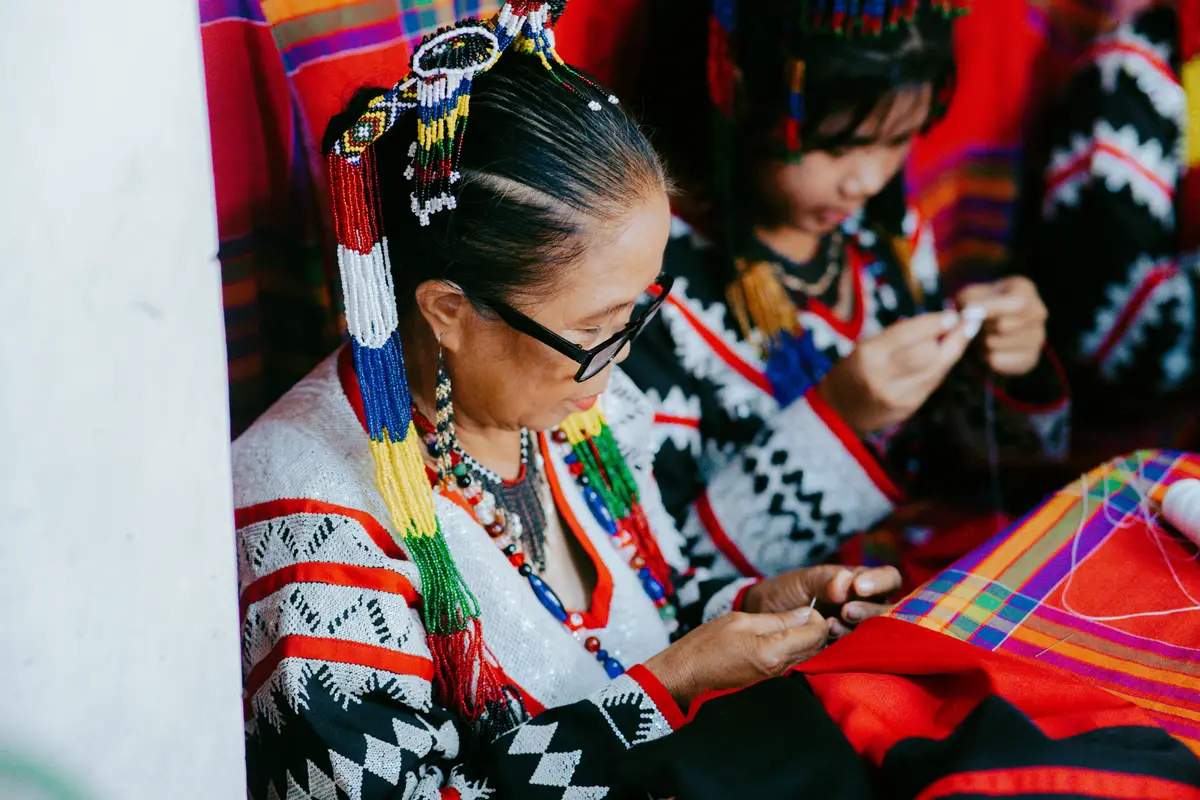 A woman and a girl in Tboli dress and beaded headpieces threading beads