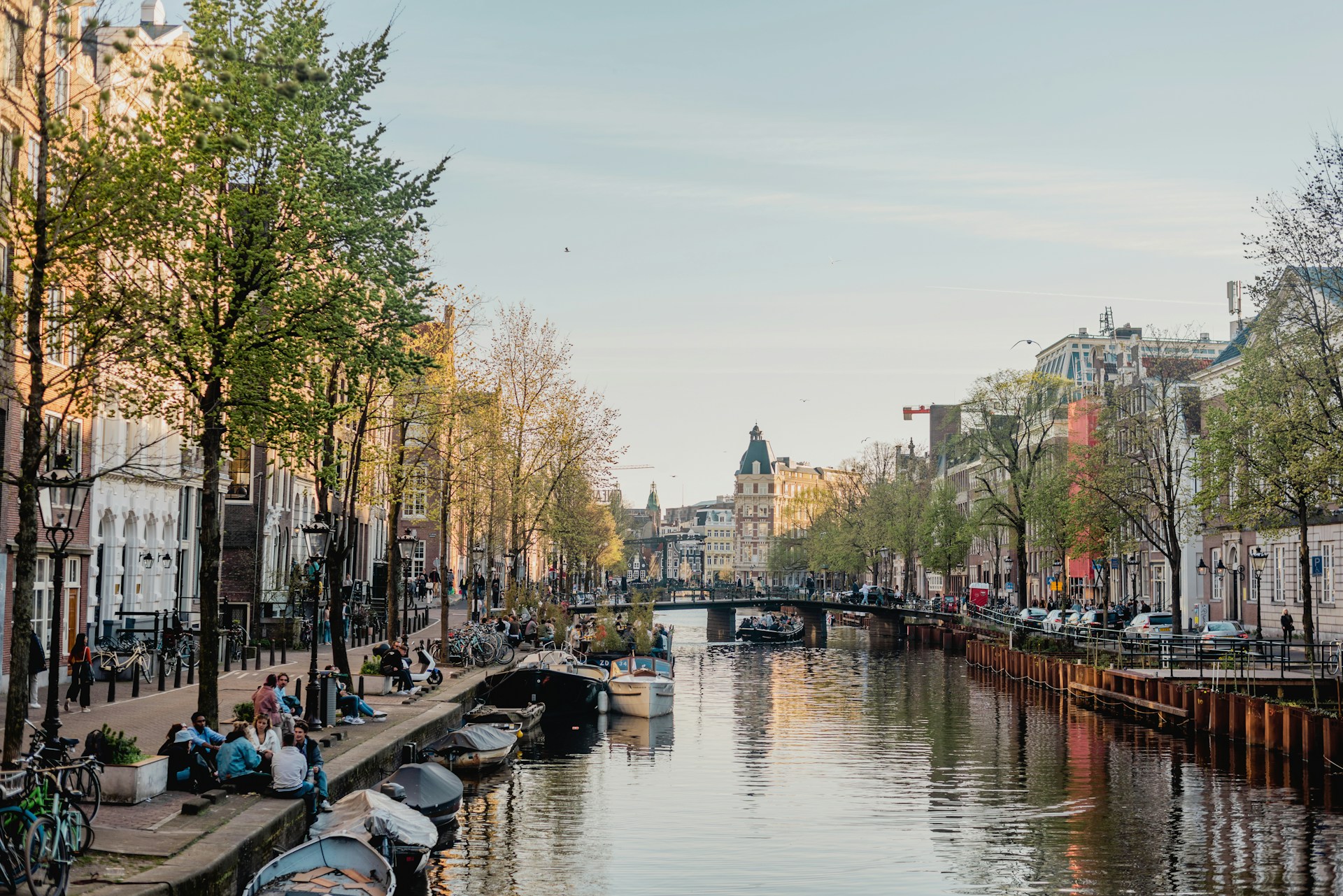 Scene of a calm canal in Amsterdam lined with boats, trees, and people sitting along the edge, with traditional Dutch buildings and a bridge in the background under a clear sky.