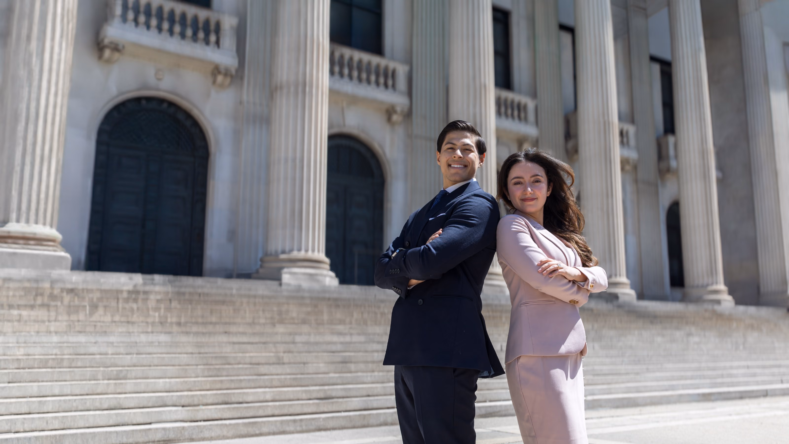 A man in a dark suit and a woman in a light pink suit stand back to back with arms crossed outside a large building with tall columns and steps.