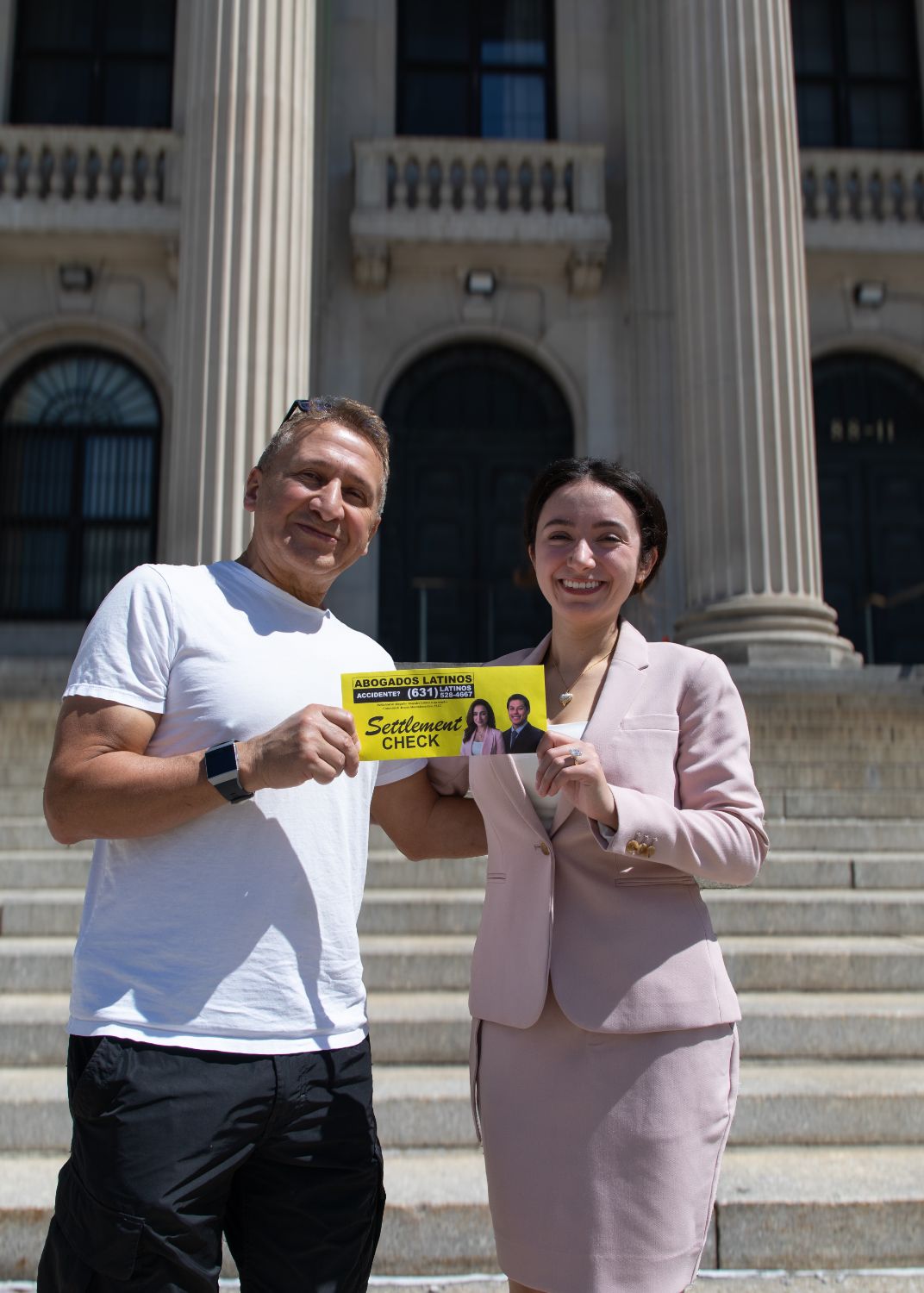 A smiling man in a white t-shirt and black pants stands next to a smiling woman in a light pink business suit on outdoor steps of a building with large columns, both holding a yellow sign that reads 'Abogados Latinos Accidente? (631) 528-4667 Settlement Check' with small photos of a man and woman on the sign.
