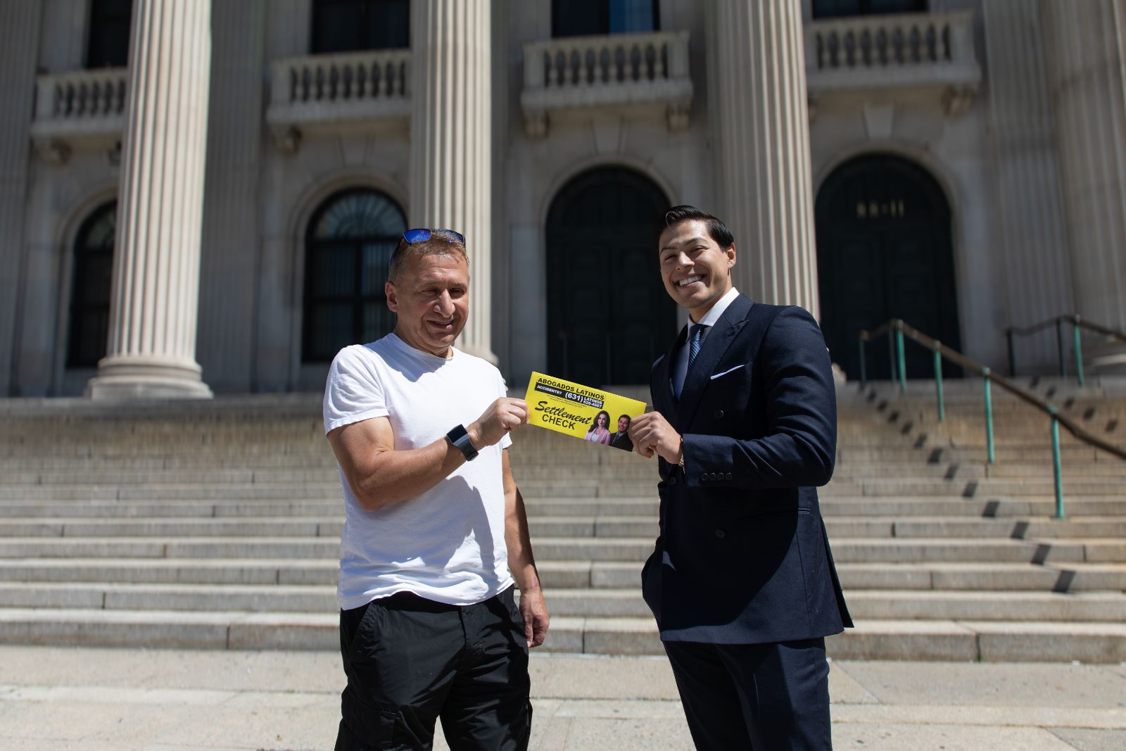 Two men standing on steps in front of a large building with columns, smiling and holding a yellow card that reads 'Abogados Latinos' and 'Settlement Check'. One man wears a white t-shirt and black pants with sunglasses on his head, and the other wears a dark suit and tie.