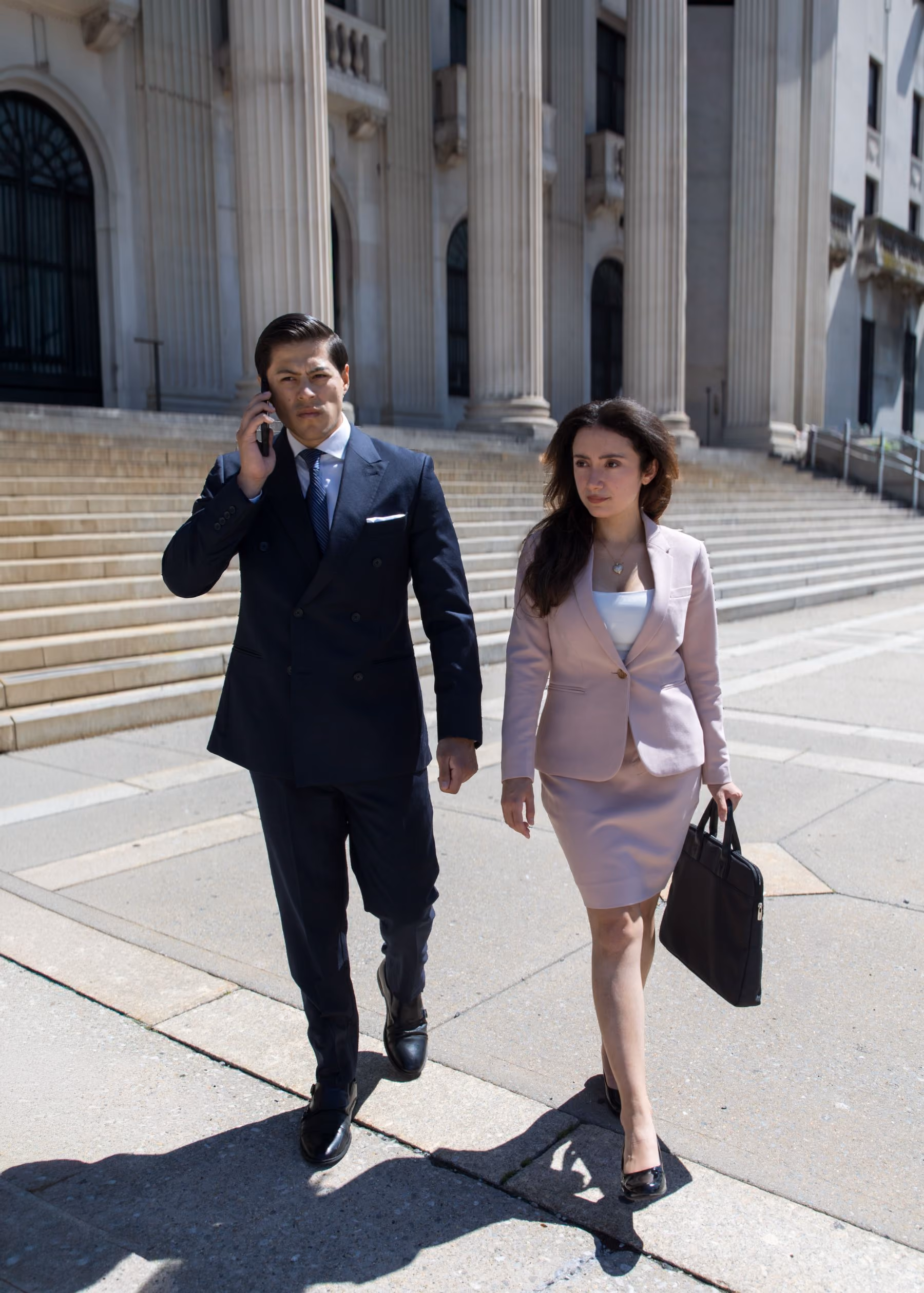 A man in a dark suit talking on a cellphone and a woman in a light pink skirt suit carrying a black briefcase walking outside in front of a building with large columns and steps.