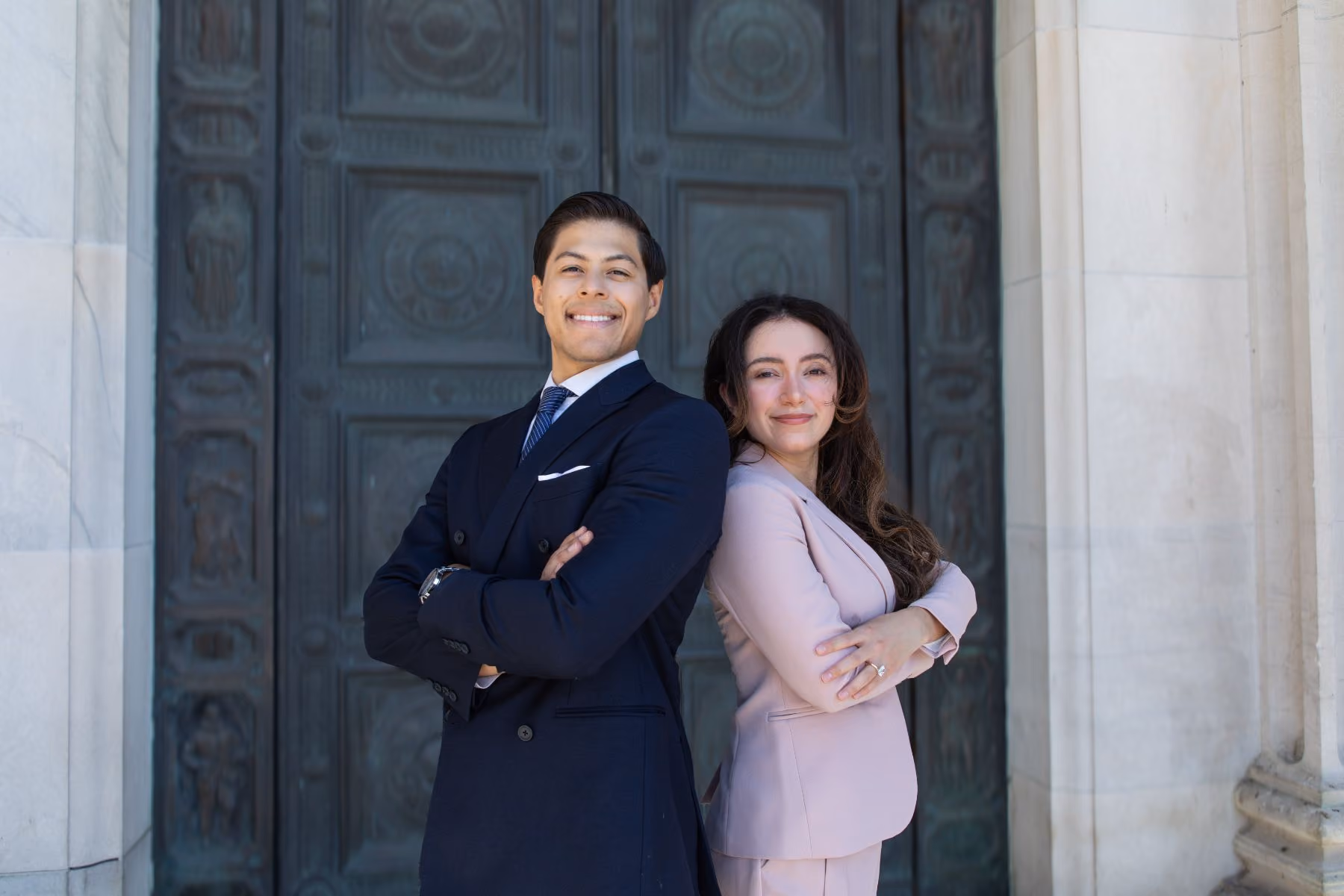 A confident young man and woman standing back to back with arms crossed in front of a large ornate door, both dressed in business attire and smiling.