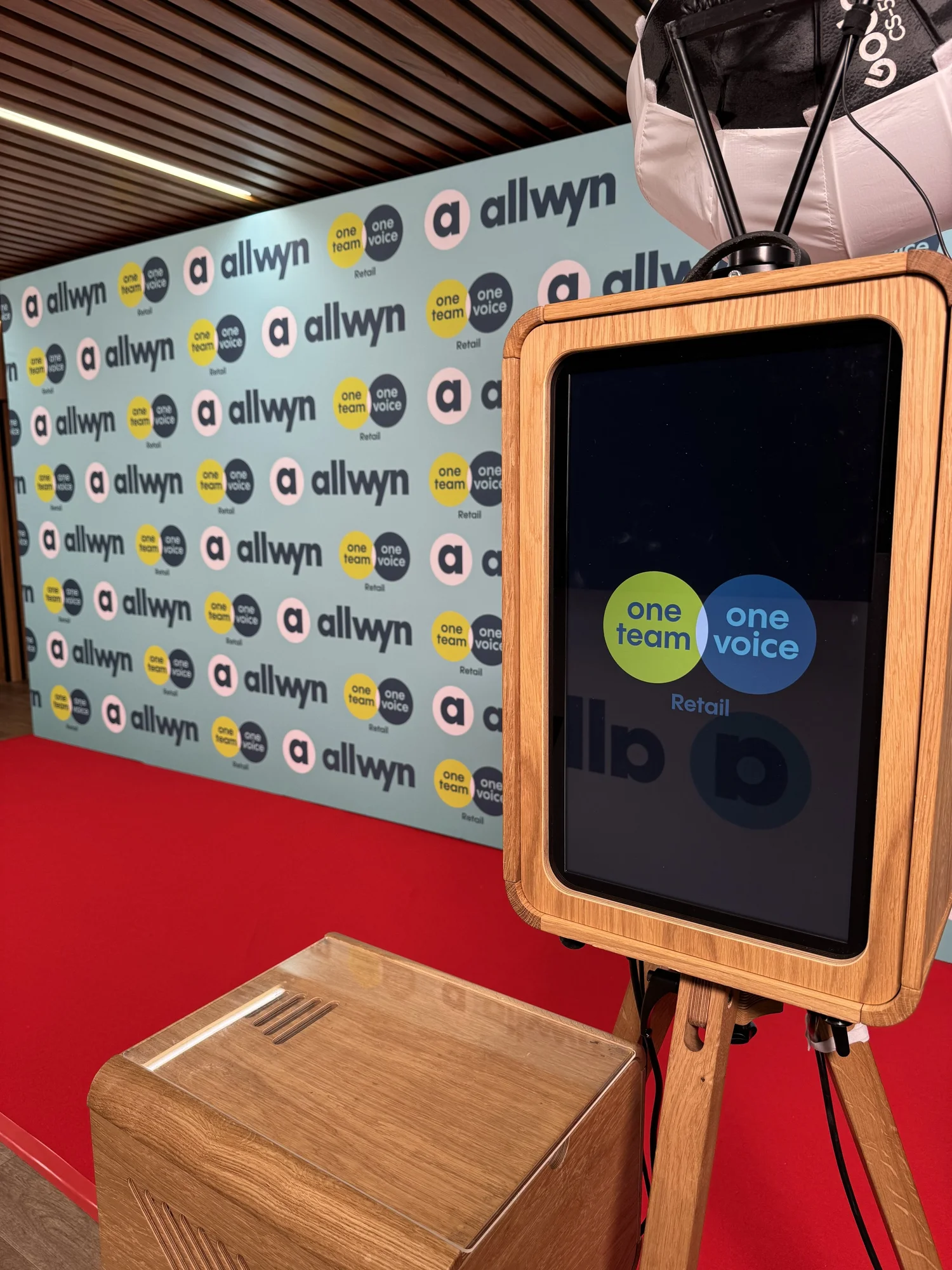 A wooden kiosk with a large touchscreen displaying the logos of 'one team' and 'one voice' in front of a light blue wall with a repeating pattern of 'Allwyn' logos and slogans 'one team' and 'one voic