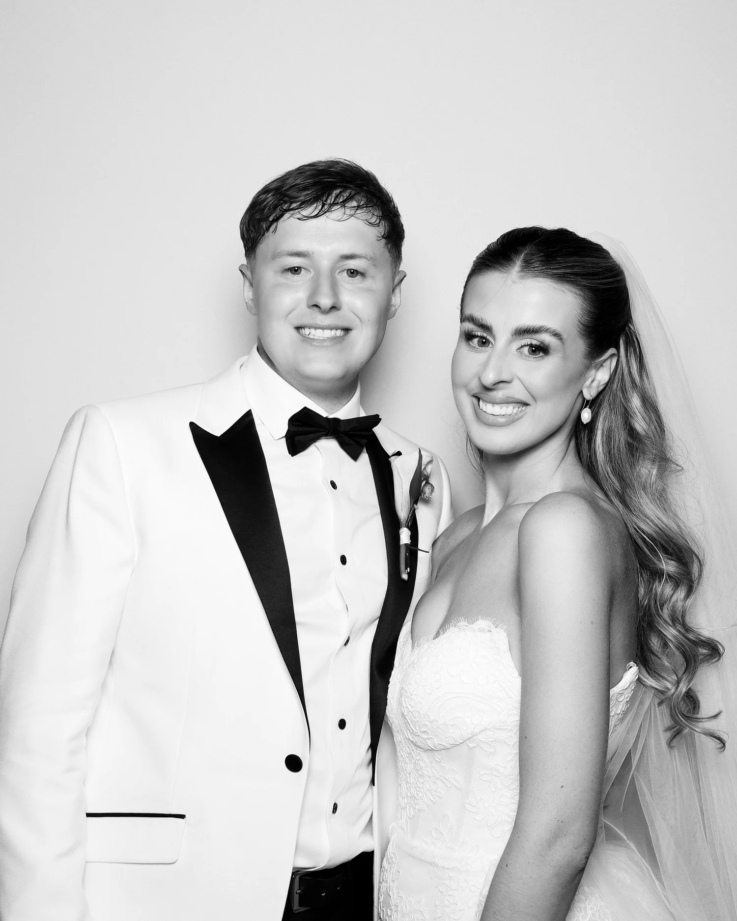 A black and white photo of a couple, dressed in formal attire, smiling and looking at the camera. The man is wearing a tuxedo with a bow tie, and the woman is in a strapless dress with earrings, with