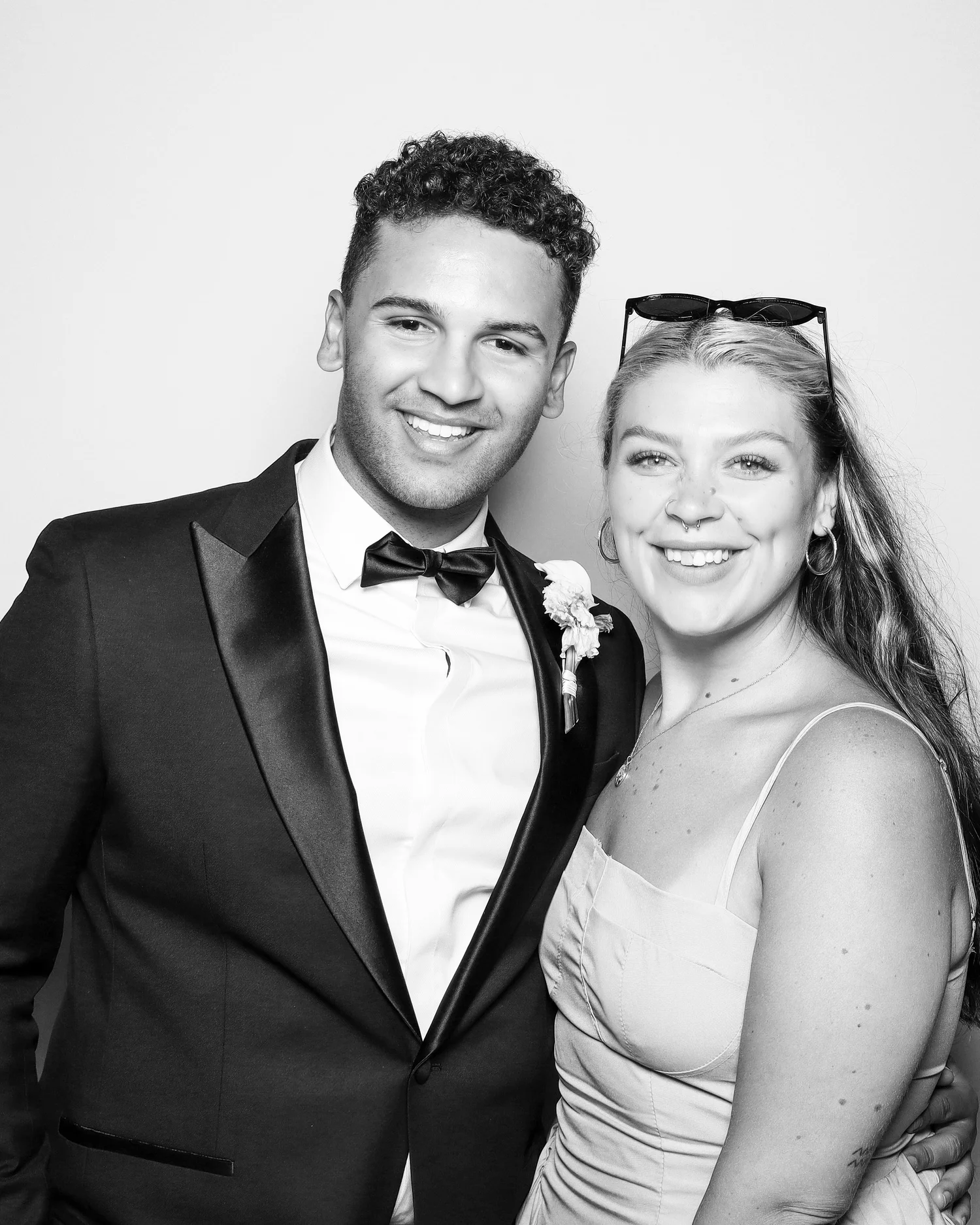 A black and white photo of a man and a woman close together, smiling at the camera. The man is dressed in a tuxedo with a bow tie, and the woman has sunglasses on her head, a septum pierced nose, and