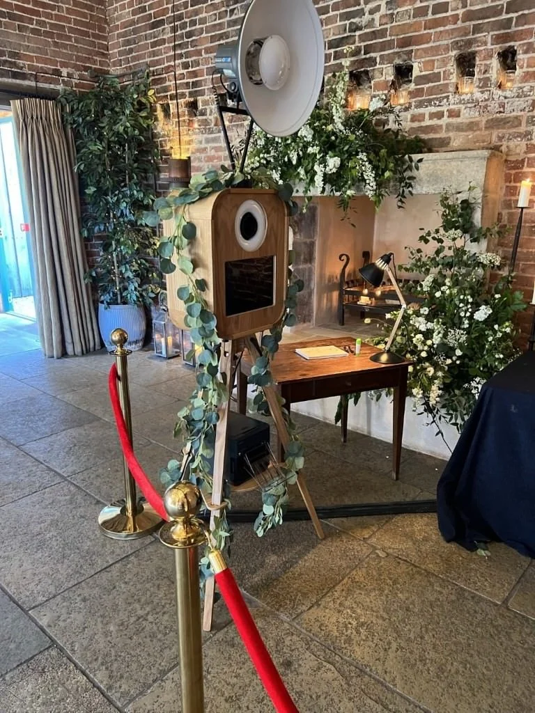 Vintage-style photo booth setup with camera, tripod, and lighting in a rustic, brick-walled venue; decorated with greenery and flowers; surrounded by a red rope barrier.