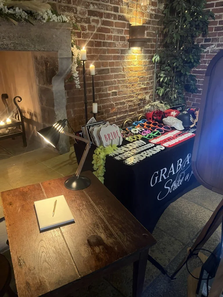 Photo booth setup with props, brick backdrop, and table with guest book and pen.