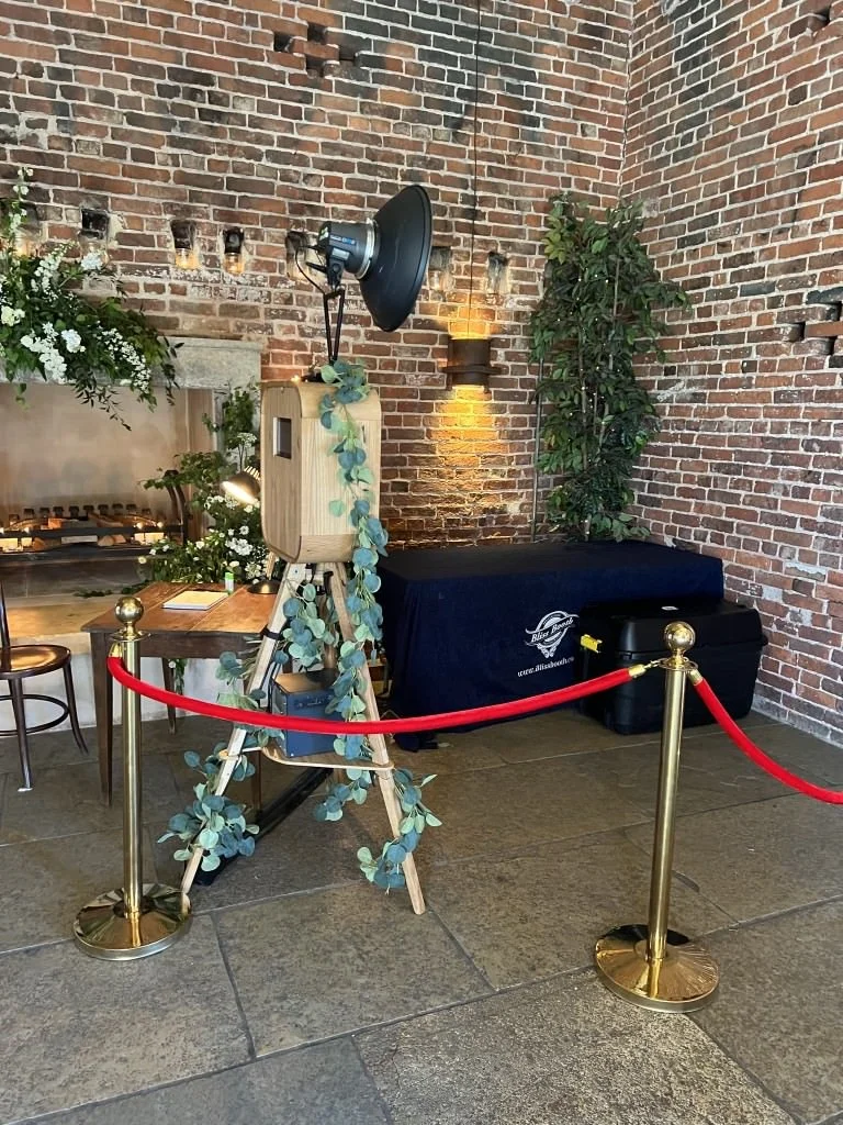 Vintage photo booth with a rustic design and greenery decoration in front of a brick wall, surrounded by a red velvet rope on golden stanchions. There are flower arrangements and chairs nearby, creating a cozy, decorated atmosphere.