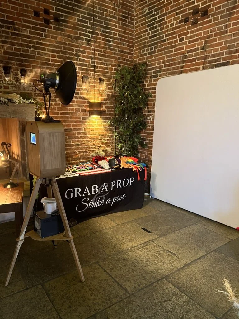 Photobooth setup with props in a room with brick walls, featuring a vintage-style camera setup, a table with colorful accessories, and a white backdrop.
