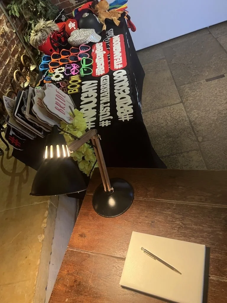 A table display with various party props, including colorful sunglasses, hats, and masks, alongside hashtag signs related to weddings. A nearby desk features a black lamp and a closed notebook with a silver pen on top.