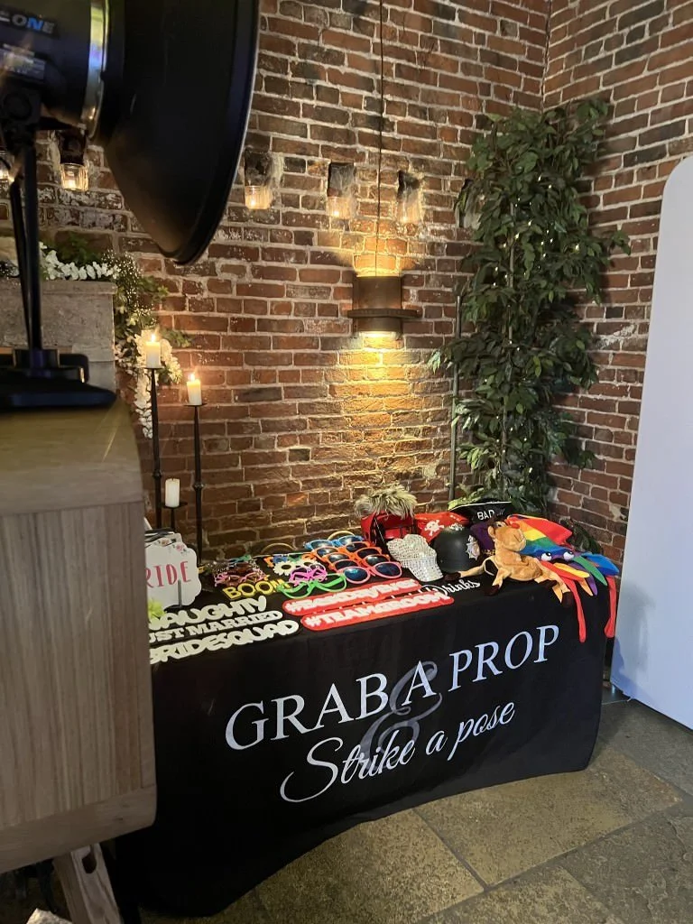 Photo booth setup with props including colorful glasses, hats, and signs displayed on a table covered with a 'Grab a Prop & Strike a Pose' cloth, set against a brick wall background with soft lighting and plants.